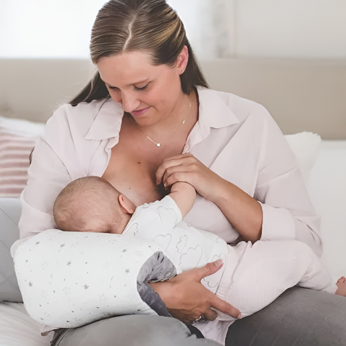 Mother breastfeeding baby with soft pillow in cozy home, baby wearing cloud-print outfit