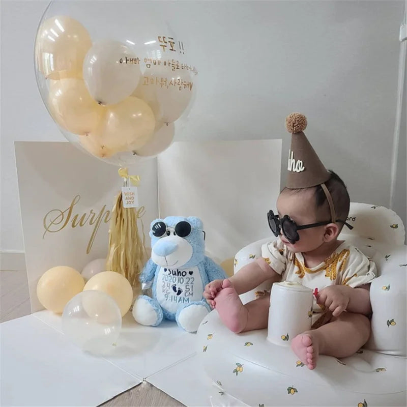 Baby in party hat and sunglasses on floral baby chair with balloons and blue teddy bear