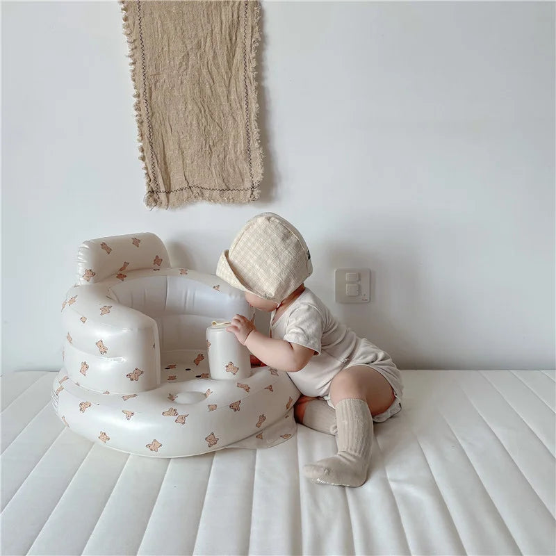 Baby wearing cream outfit and bonnet sitting next to inflatable baby chair with bear prints