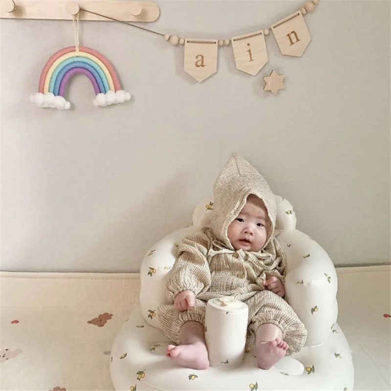 Baby in beige hooded outfit sitting in soft baby chair, rainbow wall decor, educational playroom