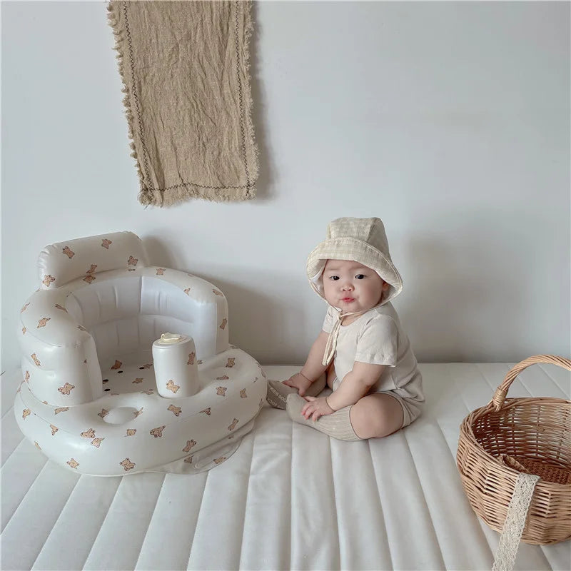 Baby in cream outfit and hat sitting on bed with inflatable chair and wicker basket, Kid Zone
