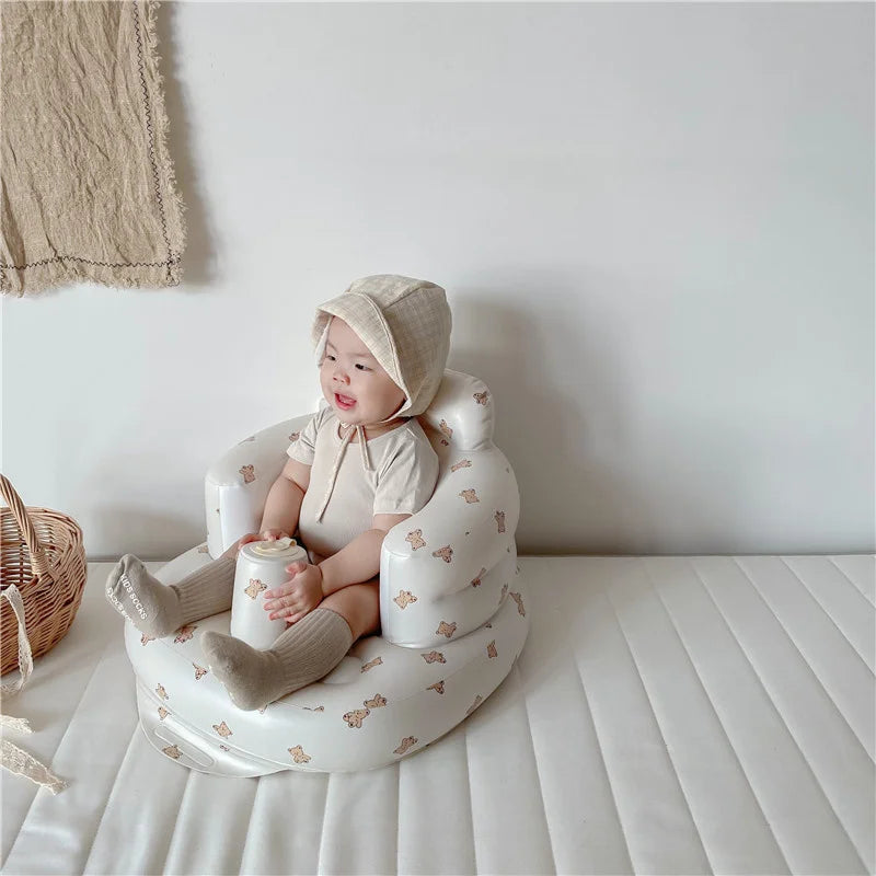 Smiling baby in beige romper and bonnet seated in a soft baby chair with teddy bear print