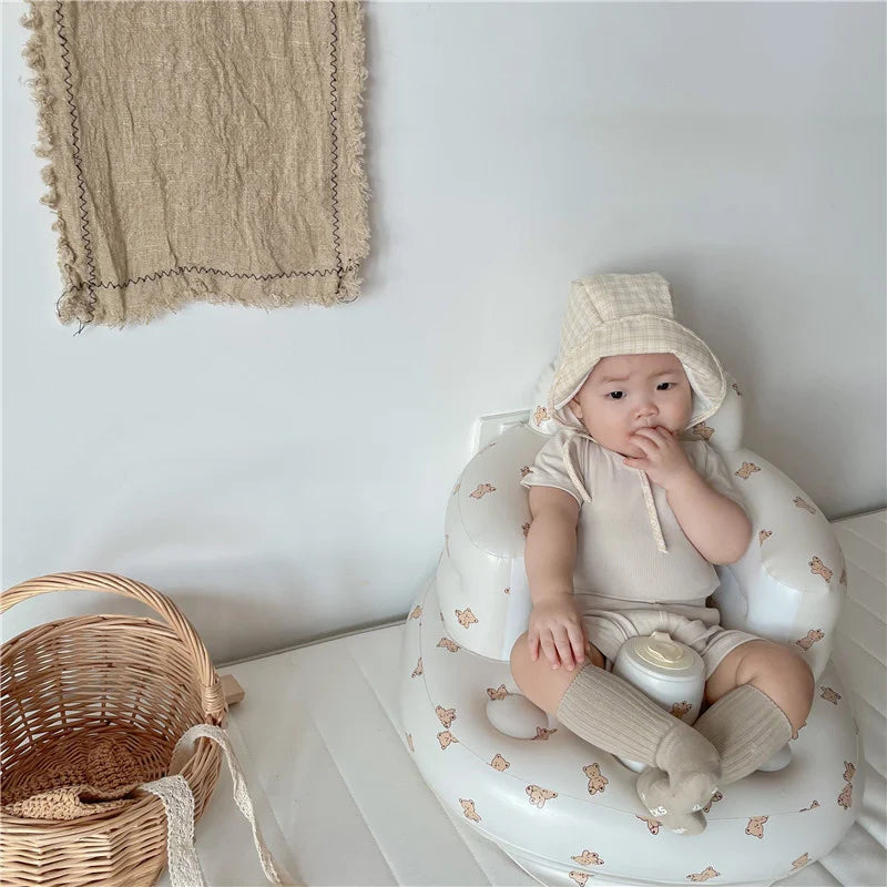 Baby in beige outfit and bonnet sitting in bear-patterned chair with knit socks, basket nearby