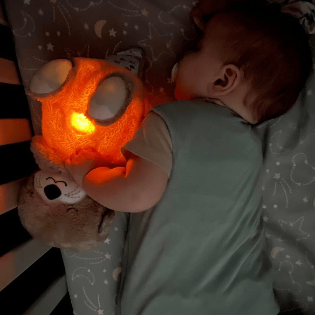 Sleeping baby cuddling glowing otter calming toy in crib, baby essentials at Kid Zone